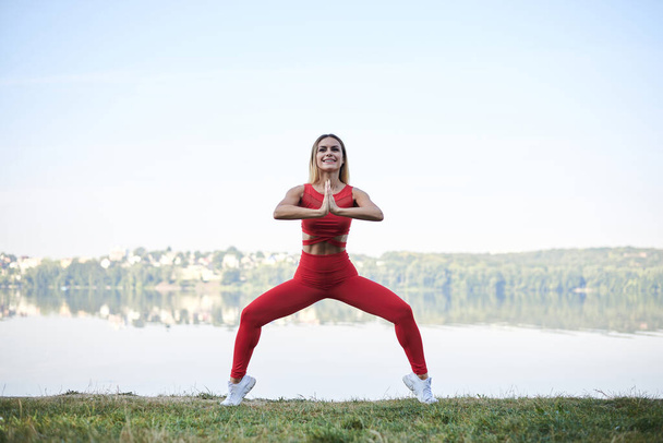 Young blond woman, wearing red leggings, top and white sneakers, doing morning exercises by lake in summer. Fitness yoga stretching training outside by water. Healthy life concept. Active way of life - Fotó, kép