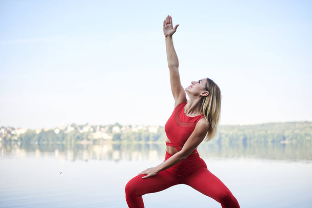 Young blond woman, wearing red leggings, top and white sneakers, doing morning exercises by lake in summer. Fitness yoga stretching training outside by water. Healthy life concept. Active way of life - Foto, Imagen