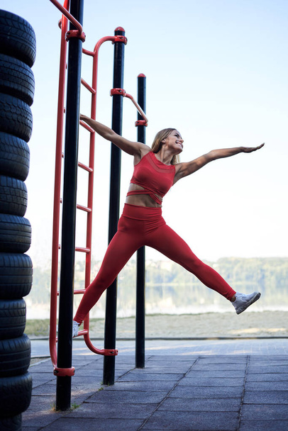 Young blond woman, wearing red sports outfit and white sneakers, doing exercises on swedish wall outside by city lake in the morning. Healthy life concept. Female fitness training - 写真・画像
