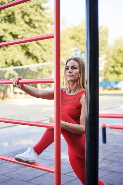 Young blond woman, wearing red sports outfit and white sneakers, doing exercises on swedish wall outside by city lake in the morning. Healthy life concept. Female fitness training - Photo, Image