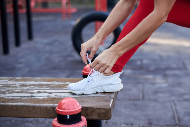 Young woman, wearing red fitness outfit, tying shoe laces on white sneakers. Process of fastening sport shoes. Close-up shot of foot and hands, wearing sneakers, preparing for training on playground. - Photo, Image