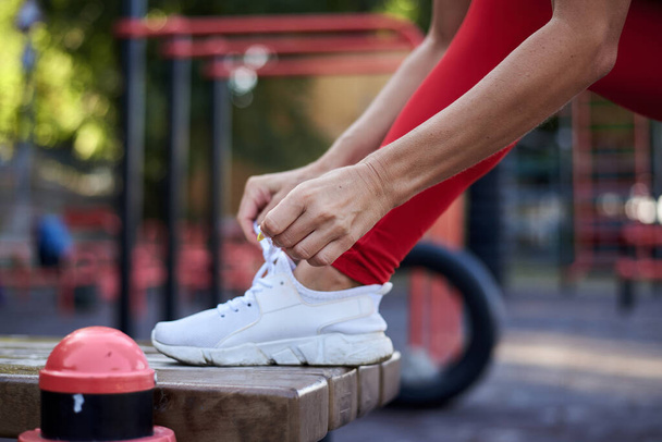 Mujer joven, vestida con traje de fitness rojo, atando cordones de zapatos en zapatillas blancas. Proceso de sujeción de zapatos deportivos. Primer plano del pie y las manos, usando zapatillas, preparándose para el entrenamiento en el patio de recreo
. - Foto, imagen