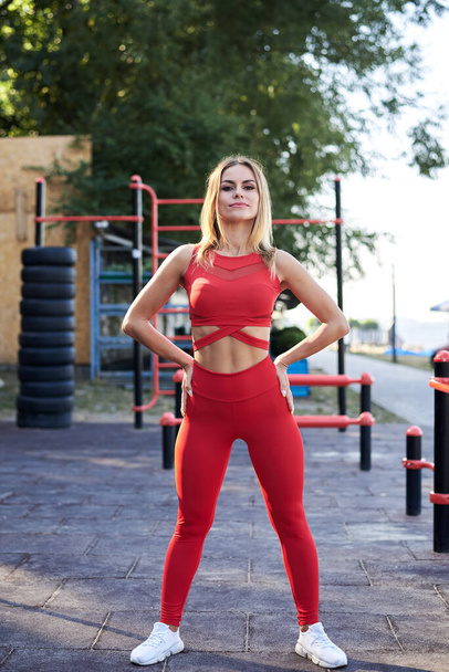 Young blond woman, wearing red fitness outfit standing on sports playground in summer morning. Healthy active life concept. Full-length portrait of girl in process of doing exercises outside in city. - Foto, Imagem