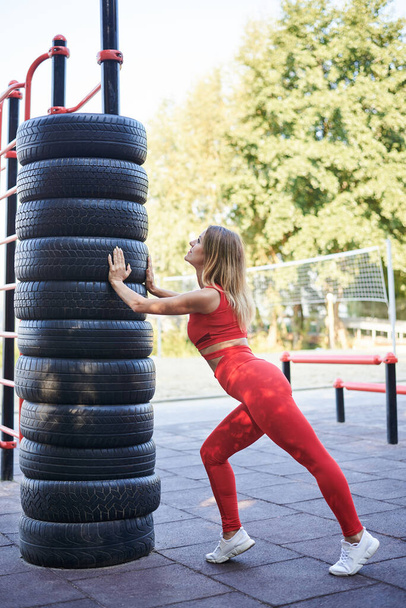 Young blond woman, wearing red fitness outfit standing on sports playground by old tires bar in summer. Healthy active life concept. Full-length portrait of girl in process training exercises outside - Fotó, kép