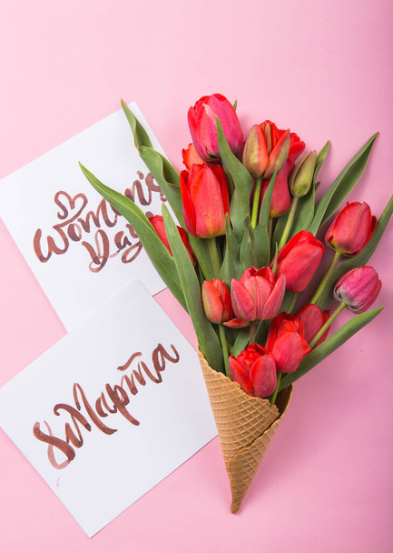 rote schöne Tulpen in einem Eis Waffelkegel mit Karte Womans Day auf einem farbigen Hintergrund. Konzeptionelle Idee eines Blumengeschenks. Frühlingsstimmung - Foto, Bild