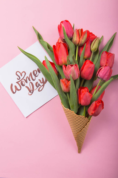 red  beautiful tulips in an ice cream waffle cone with card Womans Day on a color background. Conceptual idea of a flower gift. Spring mood - Фото, изображение