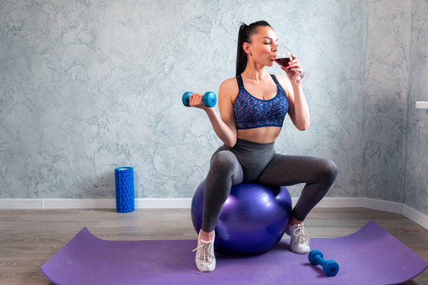 Mujer joven en traje deportivo haciendo ejercicio de fitness con mancuerna y vaso de vino tinto, sentado en la pelota de fitness en casa - Foto, Imagen