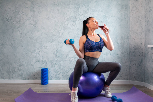 Молодая женщина в спортивном костюме занимается фитнесом с гантелями и пьет красное вино, сидит на фитнес-мяч дома - Фото, изображение