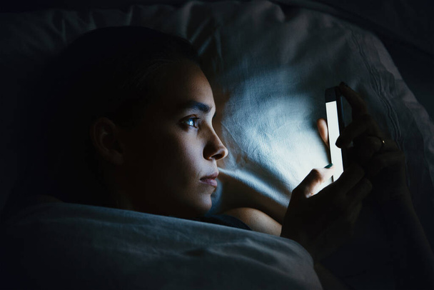 Jeune belle femme au lit en utilisant le téléphone mobile tard dans la nuit à la chambre sombre, concept de dépendance à Internet - Photo, image