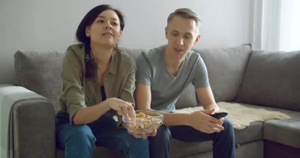 Portrait of a happy young couple eating popcorn and emotionally rooting for her favorite team while watching a sports championship on TV at home. - Materiaali, video