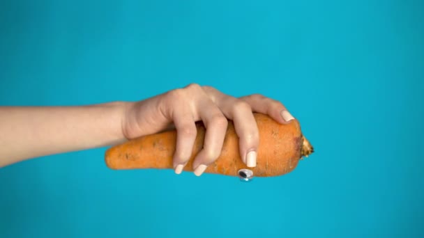 Karotte mit Augen in einer Frauenhand aus nächster Nähe. Zuckerbrot sieht sich auf blauem Grund um. Vertikales Video. - Filmmaterial, Video