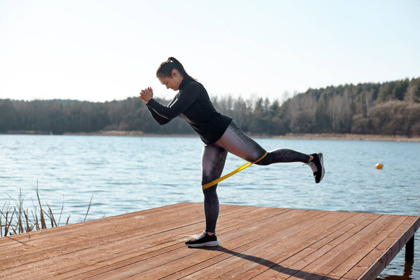 Zdravá mladá žena provádí cvičení s fitness elastickými stuhami na molu na břehu jezera. Koncept zdravého životního stylu. Boční pohled - Fotografie, Obrázek