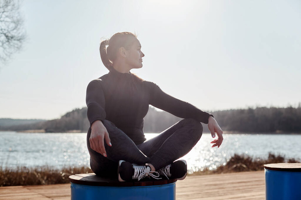 Молодая привлекательная девушка в спортивной одежде сидит в позе Лотоса на бочке на берегу озера. Девушка медитирует после тренировки. Концепция здорового образа жизни
 - Фото, изображение