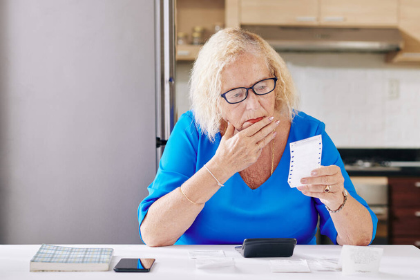 Θλιβερή ηλικιωμένη γυναίκα τρίβει το πηγούνι της όταν ελέγχει λογαριασμούς - Φωτογραφία, εικόνα