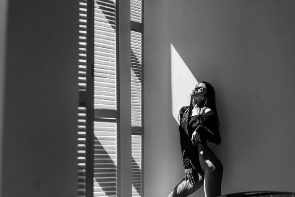 Young beautiful girl model, tall, in a black body, in a large bright room, bright sunlight from the window falls on her - Фото, изображение