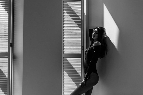 Молодая красивая девушка-модель, высокая, в черном теле, в большой светлой комнате, на нее падает яркий солнечный свет из окна - Фото, изображение