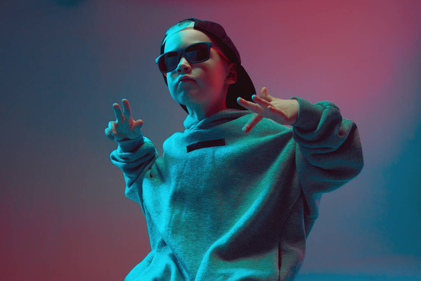Portrait of a cool boy child in a rap image, stylishly posing in a hoodie, sunglasses and a cap on a neon background. - Foto, imagen