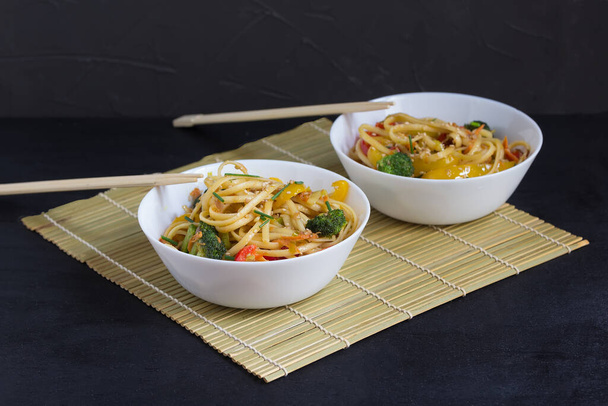 Comida asiática, duas taças de macarrão com legumes e molho de soja em um tapete de bambu, em um fundo preto
 - Foto, Imagem