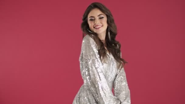 Egy flörtölő mosolygós nő fényes flitteres ruhában pózol, és küld egy légcsókot elszigetelt rózsaszín fal háttér - Felvétel, videó