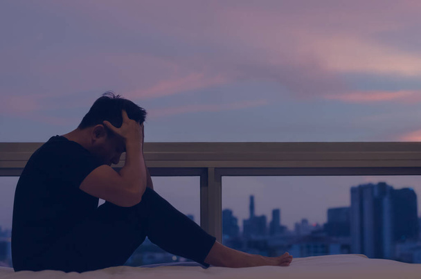 Людина, яка сидить і відчуває себе пригніченою на ліжку з видом на місто в сутінковий момент. Залишайтеся вдома, депресія і самотність концепція
. - Фото, зображення