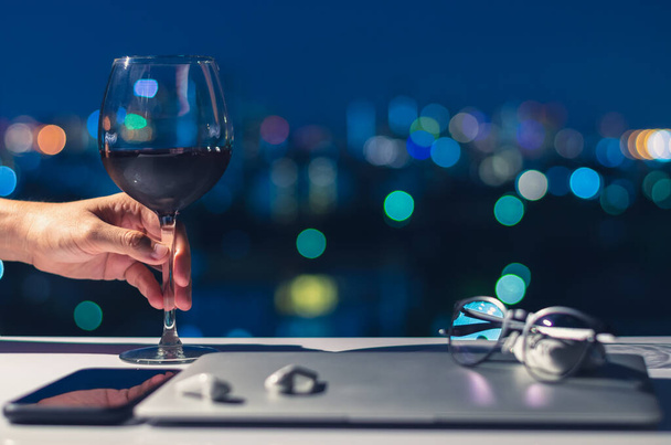 Hand houden van een glas rode wijn op tafel om te genieten van de nacht na het uitschakelen van laptop, smartphone en oortelefoon voor digitale detox, sociale ontkoppeling, gadget verslaving concept. - Foto, afbeelding