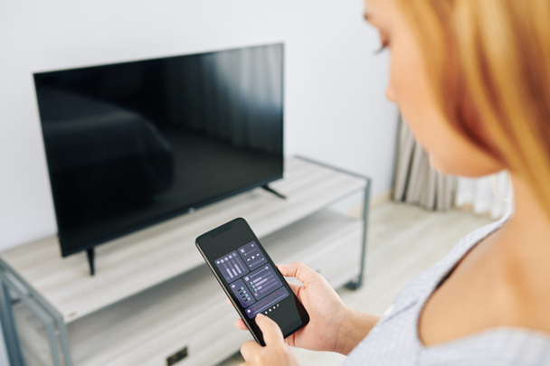 Akıllı telefondan TV uygulaması ile TV ses ayarlarını kontrol eden kadın - Fotoğraf, Görsel