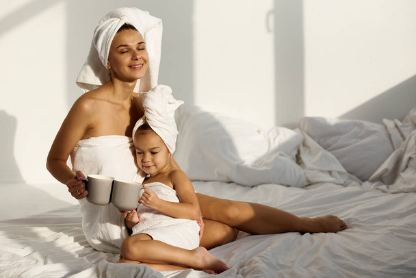 A young caucasian woman with his daughter on a bed at home in the morning, wearing in towel play and drink a milk - Fotó, kép