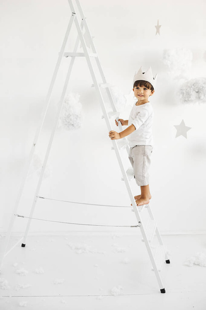 portrait of a cute little boy in crown on white ladder among clouds on a white background - Φωτογραφία, εικόνα