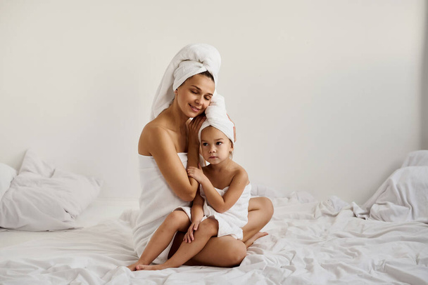 A young mother and her little daughter in white towels in the bedroom in the morning. Happy family at home.  - Foto, afbeelding