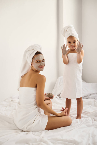 A young caucasian mother and little daughter with wrapped hair in white bath towels apply a clay mask on the mother faces - Zdjęcie, obraz