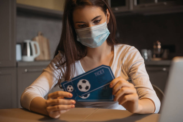 selective focus of sad woman in medical mask holding sport match tickets at home - Foto, Imagem