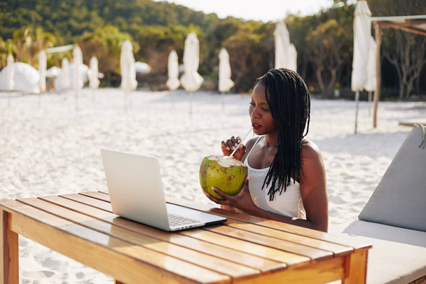 Bella giovane donna nera con dreadlocks bere cocktail di cocco e guardare webinar sul computer portatile quando si siede a tavola all'aperto
 - Foto, immagini