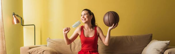 πανοραμική λήψη της ευτυχούς αθλήτριας αγγίζοντας ιατρική μάσκα και κρατώντας μπάσκετ στο σαλόνι, τέλος της έννοιας καραντίνα  - Φωτογραφία, εικόνα