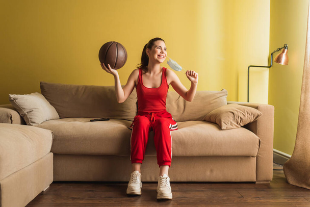 happy sportswoman touching medical mask and holding basketball in living room, end of quarantine concept  - Foto, afbeelding