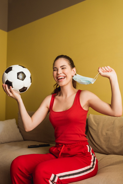 cheerful sportswoman touching medical mask and holding football, end of quarantine concept  - Φωτογραφία, εικόνα
