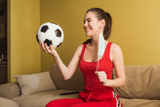 happy sportswoman touching medical mask and holding football, end of quarantine concept  - Φωτογραφία, εικόνα
