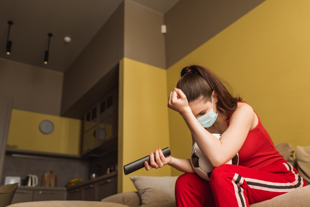 upset sportswoman in medical mask holding remote controller near football while watching championship in living room  - Fotografie, Obrázek