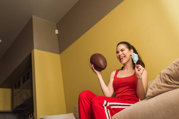 cheerful sportswoman touching medical mask and holding american football, end of quarantine concept  - Foto, Imagen