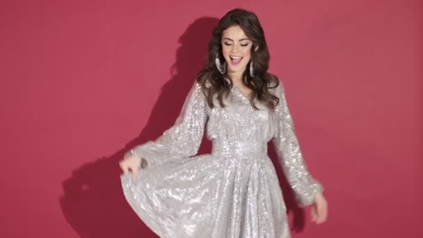 A happy smiling young woman in bright sequins dress is posing isolated over pink wall background - Materiał filmowy, wideo