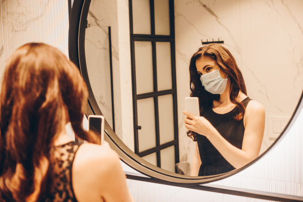 selective focus of woman in black dress and medical mask taking photo in bathroom - Photo, Image