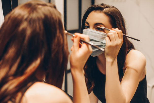 selective focus of young woman in medical mask and black dress applying eye shadow in bathroom - Photo, Image