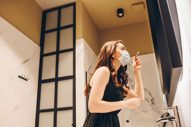 Tıbbi maskeli ve siyah elbiseli bir kadının banyoda göz farı ile kozmetik fırça tutuşu. - Fotoğraf, Görsel
