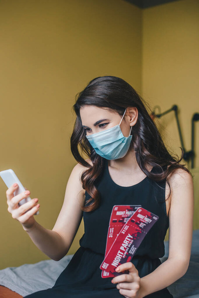 selective focus of girl in medical mask holding night party tickets and using smartphone, end of quarantine concept  - Фото, изображение