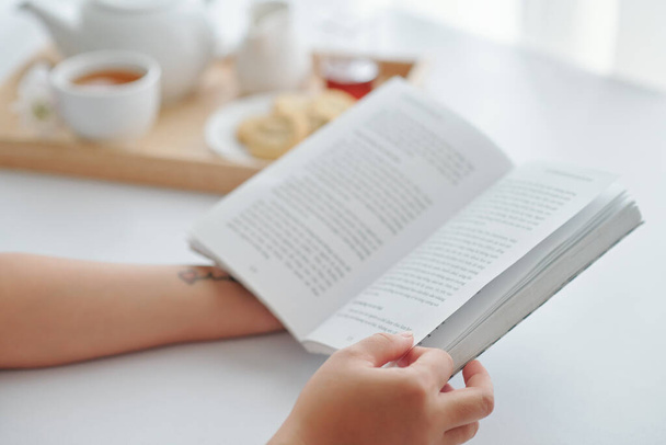 Κοντινό πλάνο εικόνα της νεαρής γυναίκας να παίρνει πρωινό και να διαβάζει βιβλίο στο σπίτι το πρωί - Φωτογραφία, εικόνα
