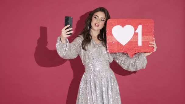 A beautiful young woman in bright sequins dress is holding a red placard with a heart on it while taking selfie isolated over pink wall background - Video