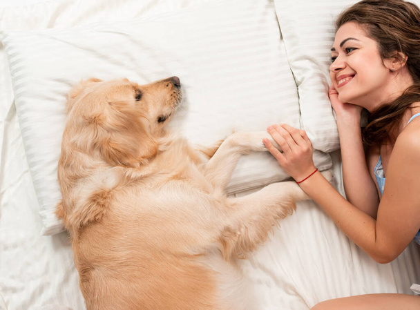 Yatakta Golden Retriever köpeğiyle pijamalarıyla oynayan neşeli kadın. İnsan ve köpek arasındaki romantik ilişki. - Fotoğraf, Görsel
