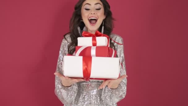 A surprised young woman in bright sequins dress is holding presents isolated over pink wall background - Video