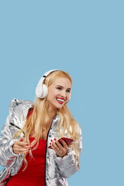 Portrait of cheerful young woman wearing casual silver jacket isolated over blue background, listening to music with headphones, dancing. Wow and surprised face - 写真・画像