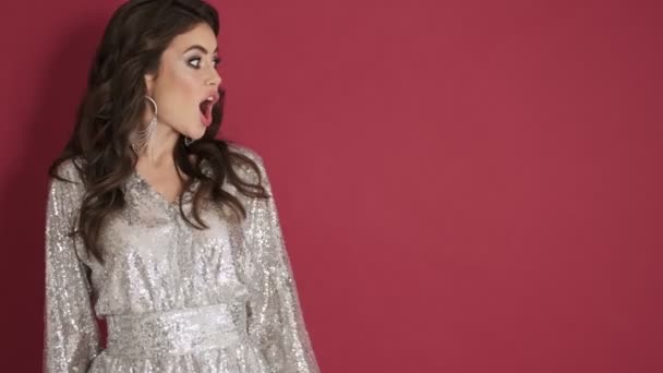 A surprised smiling young woman in bright sequins dress is pointing to the side isolated over pink wall background - Metraje, vídeo