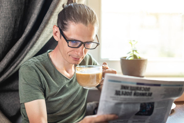 Egy jóképű fiatalember érdekes híreket olvas egy újságban, és cappuccino kávét iszik az ablak mellett. A reggel otthon töltésének koncepciója - Fotó, kép